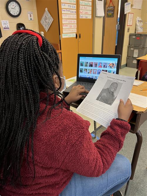 Photo shows back of female student sitting at desk using laptop to research Kehinde WIley. She's holding a printout.